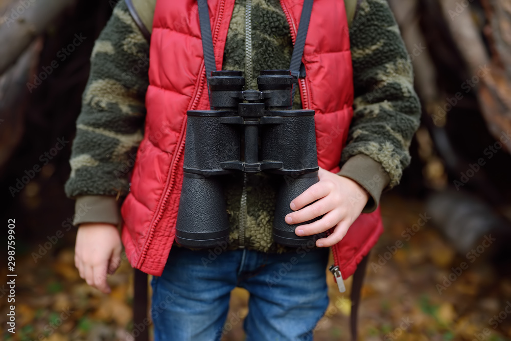 Little boy scout with binoculars during hiking in autumn forest. Behind the child is teepee hut.