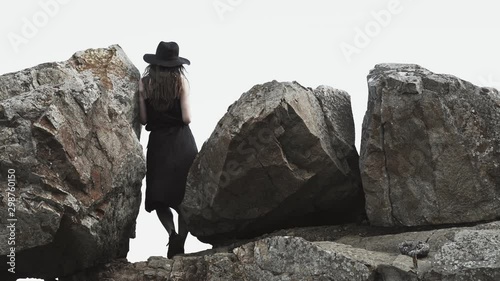 Girl in a black silk dress and a hat walking around sea cost. Ocean views and big rocks are at a background. Summer vacation, fashion. Big hat. Inspiration and relax on a shore. Landscape and nature. 