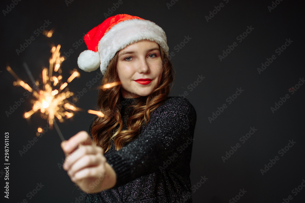 Cute girl in Santa's hat rejoices Christmas and New Year on a gray background. The girl holds a sparkler. Cute girl in Santa's hat rejoices Christmas and New Year on a gray background. The girl holds a sparkler.
