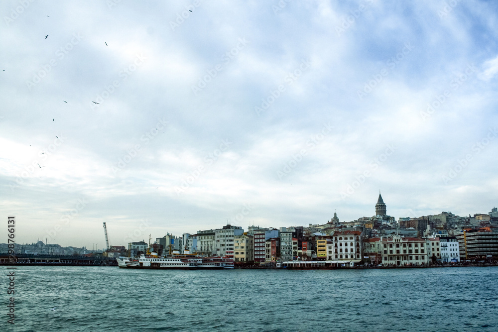 Galata Tower, on its hill, in Karakoy and Beyoglu district, taken during a cloudy winter afternoon, while the sea and ships can be visible in foreground