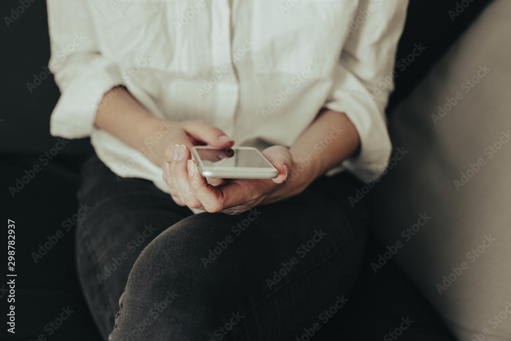 Woman hand holding white mobile phone and sitting on sofa at home.