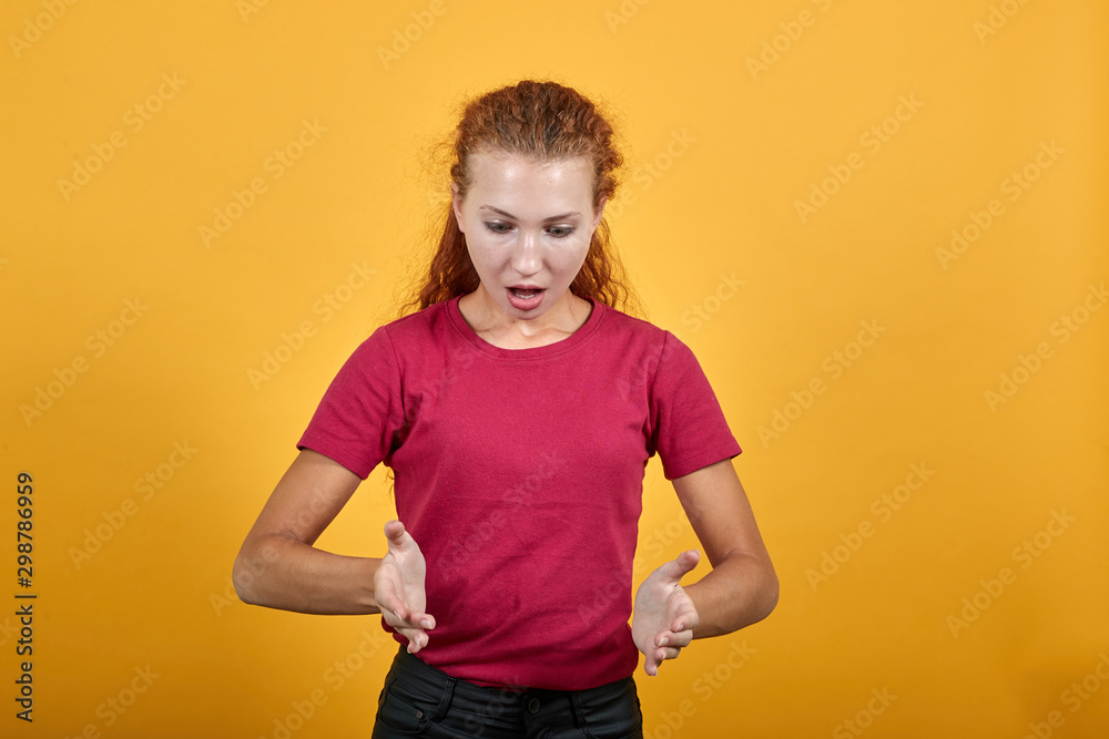 Beautiful European woman as if holding an imaginary product. Pretty girl wearing pinky clothes standing over isolated yellow background. Lady with wavy hair having eyes of gray colour. Beautiful European woman as if holding an imaginary product. Pretty girl wearing pinky clothes standing over isolated yellow background. Lady with wavy hair having eyes of gray colour.
