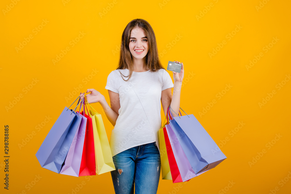 happy smiling beautiful brunette woman with colorful paper shopping bags and credit card wearing jeans and shirt isolated over yellow