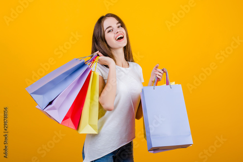 happy smiling beautiful brunette woman with colorful paper shopping bags wearing jeans and shirt isolated over yellow
