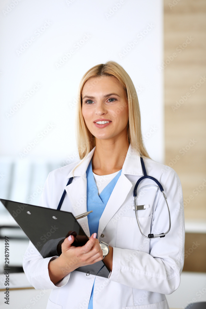Doctor woman at work in hospital excited and happy of her profession. Blonde physician controls medication history records and exam results while using clipboard. Medicine and healthcare concept Doctor woman at work in hospital excited and happy of her profession. Blonde physician controls medication history records and exam results while using clipboard. Medicine and healthcare concept