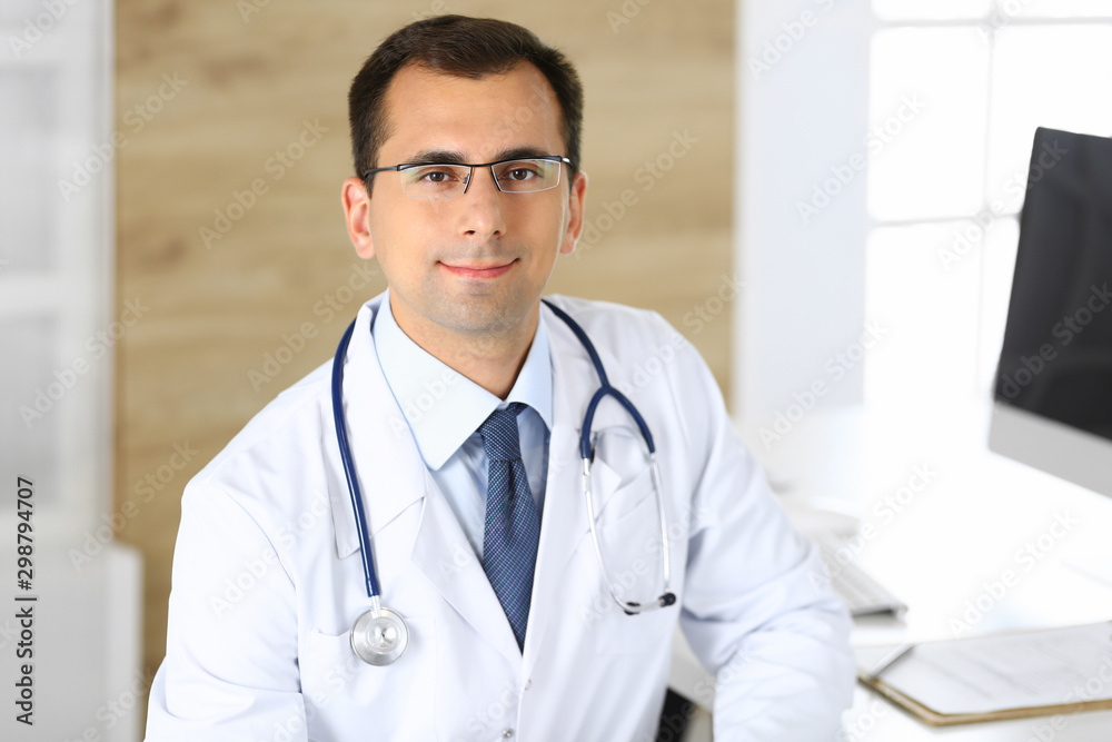 Doctor man sitting at the desk at his working place and smiling at camera. Perfect medical service in clinic. Happy future of medicine and healthcare Doctor man sitting at the desk at his working place and smiling at camera. Perfect medical service in clinic. Happy future of medicine and healthcare
