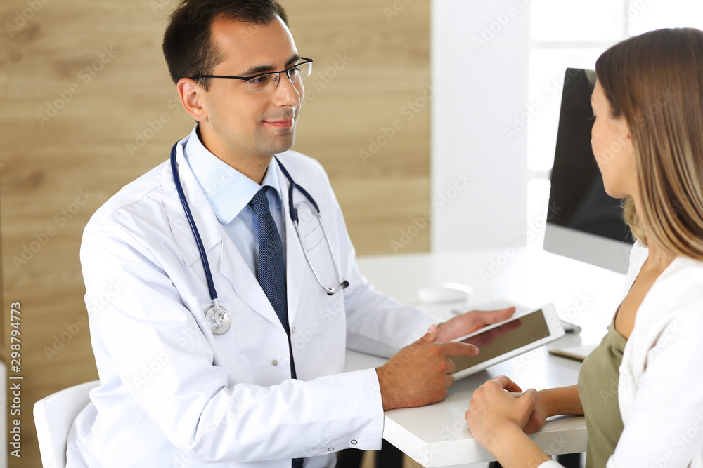Doctor and patient discussing medical exam results while sitting at the desk in clinic. Male physician using tablet computer for filling up medication history records of young woman. Data in medicine