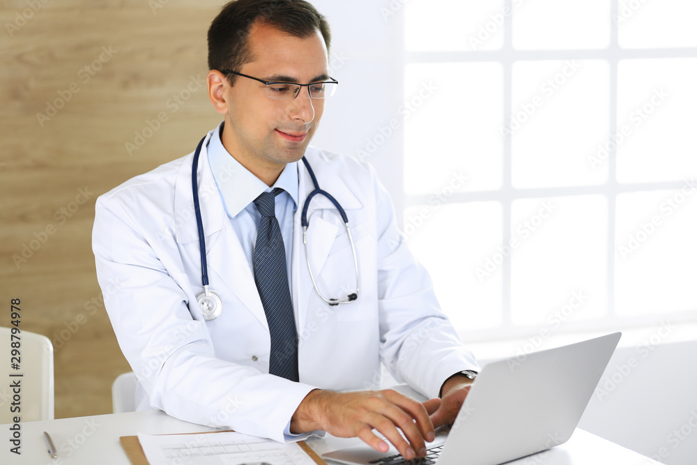 Doctor man typing at laptop computer while sitting at the desk at his working place. Perfect medical service in clinic. Data in medicine and healthcare Doctor man typing at laptop computer while sitting at the desk at his working place. Perfect medical service in clinic. Data in medicine and healthcare