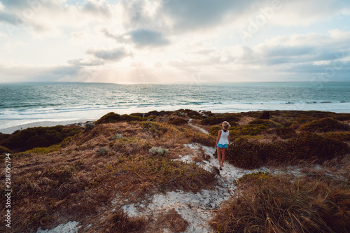 Lone woman on a windy summers evening, walking on a sand path over the beach dunes towards the beautiful ocean and sunlight coming through the clouds, at Mullaloo Beach, Perth, Western Australia. photo