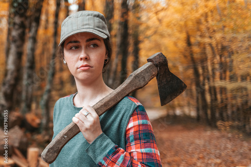 Preparation for the heating season. A young woman stands with an axe on her shoulder and looks gravely away. In the background, a pile of firewood and autumn forest. Close up