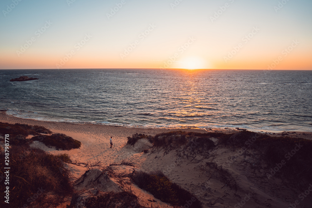 Woman walking through the sand dunes on a beach at sunset, lit by orange light at Point Peron, Rockingham, Perth, Western Australia.