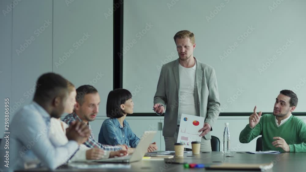 Tracking shot of diverse business team discussing market research in meeting. Man presenting document with statistical charts to colleagues in board room, business people applauding to speaker Tracking shot of diverse business team discussing market research in meeting. Man presenting document with statistical charts to colleagues in board room, business people applauding to speaker