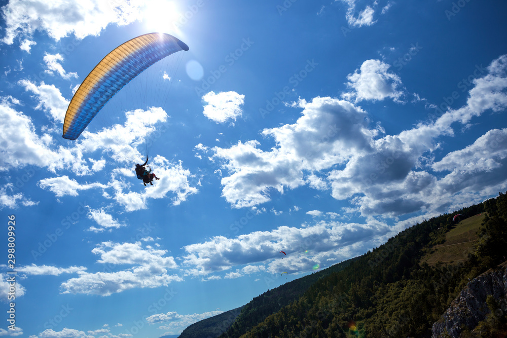 The instructor and tourist are flying high above on a yellow-blue paraglider. The sky is beautiful blue with clouds and below are conifer forests. The instructor and tourist are flying high above on a yellow-blue paraglider. The sky is beautiful blue with clouds and below are conifer forests.