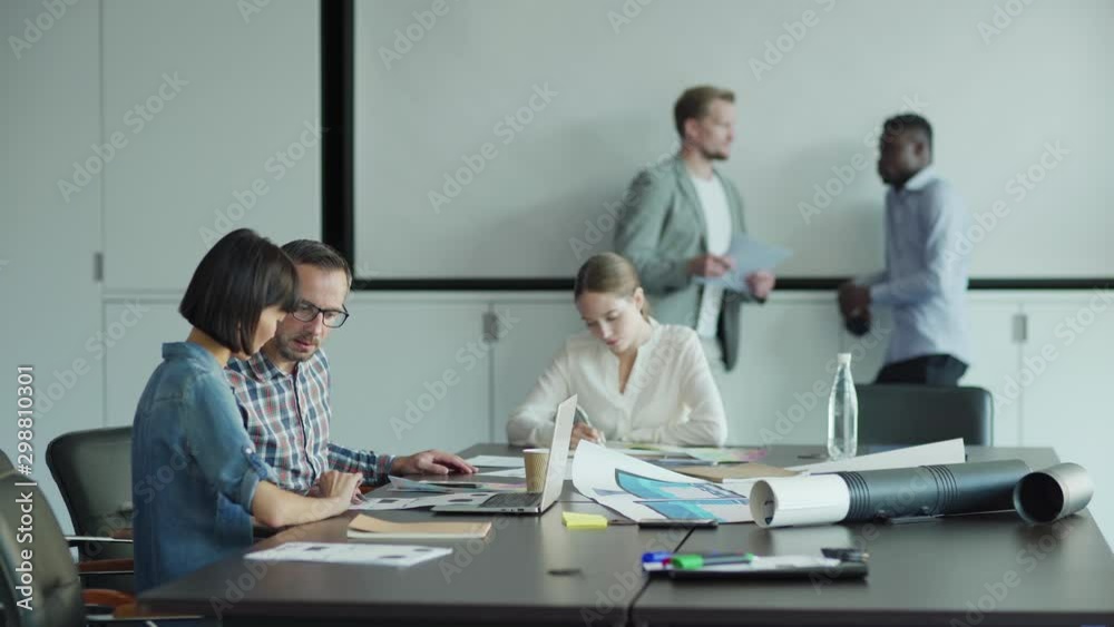 Tracking shot of business people working in office. Man and woman looking on documents and planning project, their male colleagues talking in background, young woman writing at desk nearby Tracking shot of business people working in office. Man and woman looking on documents and planning project, their male colleagues talking in background, young woman writing at desk nearby