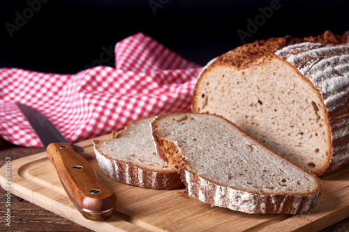 Still life with loaf and sliced wheat and rye bread with knife. On old wooden board with red cloth on black background