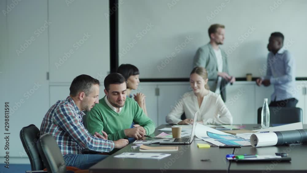 Tracking shot of diverse business people working in office meeting room. Two men discussing documents at desk, female colleagues making notes in notebook nearby. Two businessmen talking in background Tracking shot of diverse business people working in office meeting room. Two men discussing documents at desk, female colleagues making notes in notebook nearby. Two businessmen talking in background
