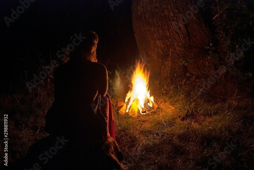 A lonely girl sits by the fire in the night forest. Bright flame of a fire.