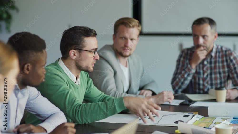 Tracking shot of diverse team of business people discussing market analysis sitting at desk in meeting. Intelligent young man in eyeglasses explaining statistics to colleagues