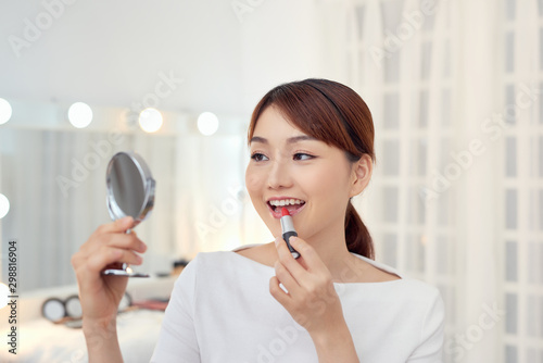  Smiling young Asian woman applying lipstick and looking to mirror.