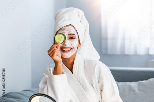 Young woman with cleansing mask on her face at home. Skin care. Woman aplying beauty mask,close up. So beautiful. Close-up of girl with beauty mask on her face looking in mirror. photo