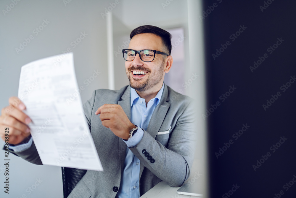 Smiling hardworking caucasian manager in gray suit sitting in modern office and reading report. Smiling hardworking caucasian manager in gray suit sitting in modern office and reading report.