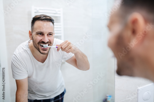 Handsome caucasian man standing in bathroom in pajamas and washing his teeth in the morning and watching himself in the mirror.