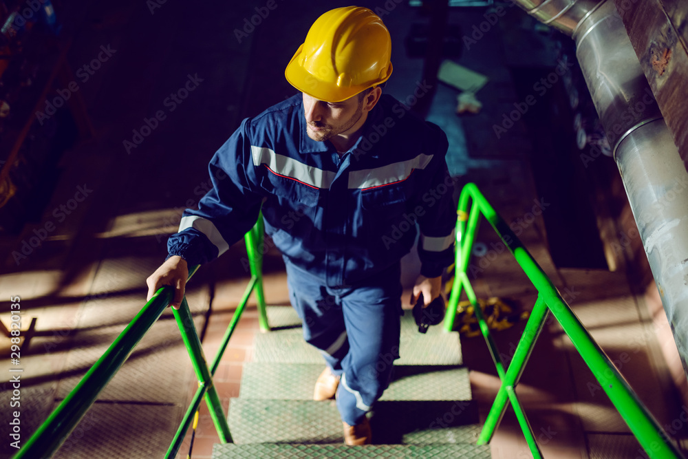 Full length of caucasian worker dressed in blue work suit and with protective helmet on head climbing up the stairs. Energy plant interior. - obrazy, fototapety, plakaty 
