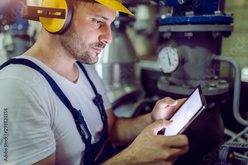 Side view of dedicates hardworking plant worker in overalls, with protective helmet on head and antiphons on ears using tablet.