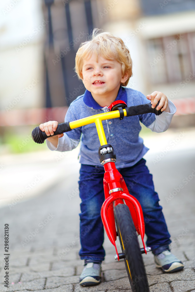 Active blond kid boy in colorful clothes driving balance and learner's bike or bicycle in domestic garden. Toddler child dreaming and having fun on warm summer day. outdoors movement game for children Active blond kid boy in colorful clothes driving balance and learner's bike or bicycle in domestic garden. Toddler child dreaming and having fun on warm summer day. outdoors movement game for children