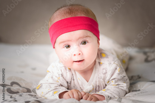 Adorable 6 months old Baby girl infant on a bed on her belly with head up looking into camera with her big eyes. Natural light.