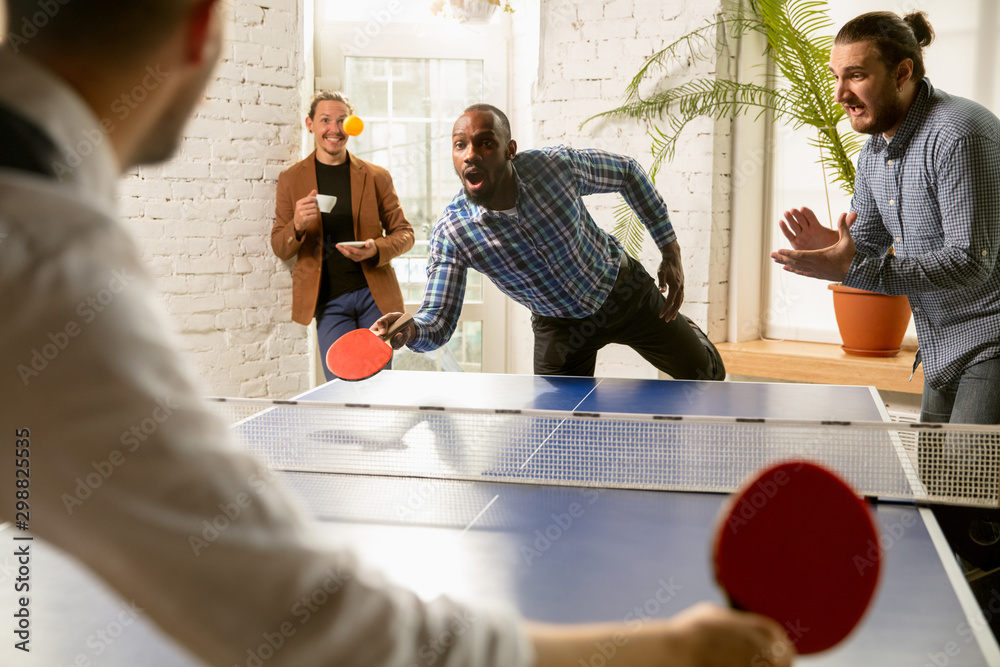 Fotka „Young people playing table tennis in workplace, having fun. Friends  in casual clothes play ping pong together at sunny day. Concept of leisure  activity, sport, friendship, teambuilding, teamwork.“ ze služby Stock