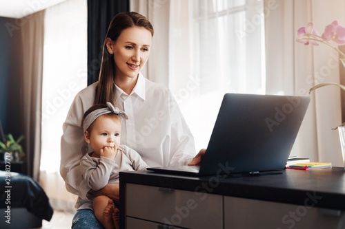 Female freelance author typing on laptop and holding baby on knees. Vacancies for home-based remote work for young parents. Distant career for professionals in digital segment with little children.