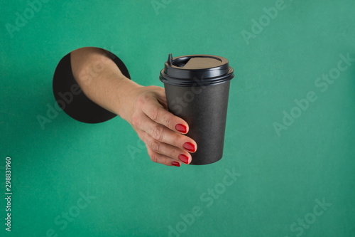 Female hand holding paper black coffee cup isolated through round hole in green paper. Concept