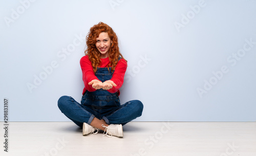 Redhead woman with overalls sitting on the floor holding copyspace imaginary on the palm to insert an ad