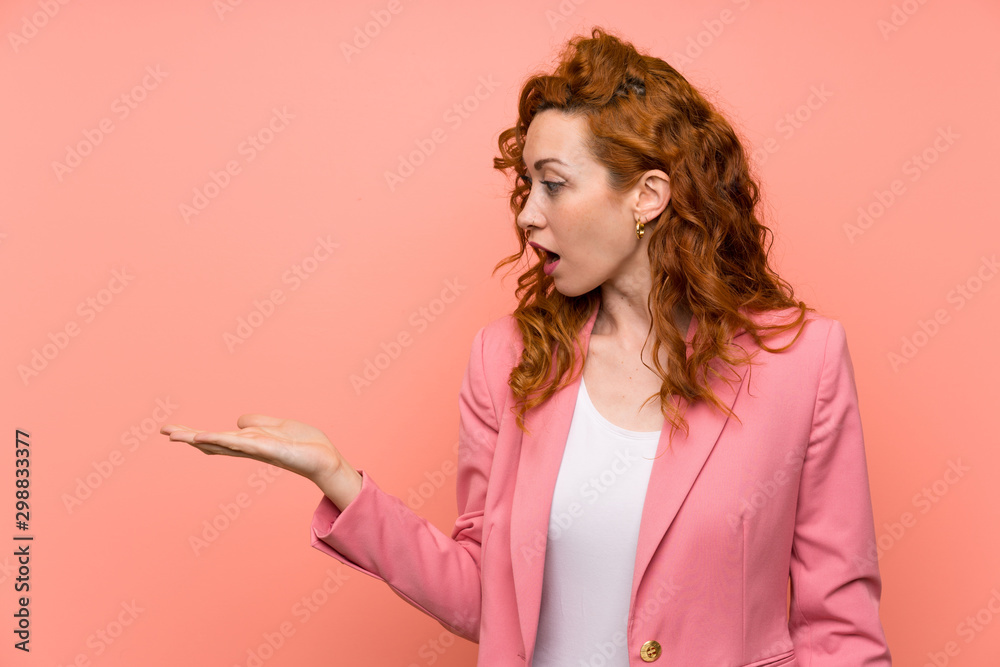 Redhead woman in suit over isolated pink wall holding copyspace imaginary on the palm Redhead woman in suit over isolated pink wall holding copyspace imaginary on the palm