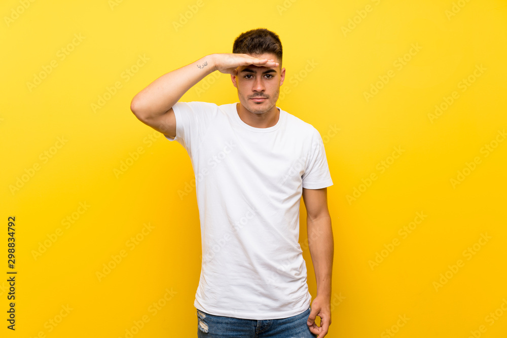 Young handsome man over isolated yellow background looking far away with hand to look something