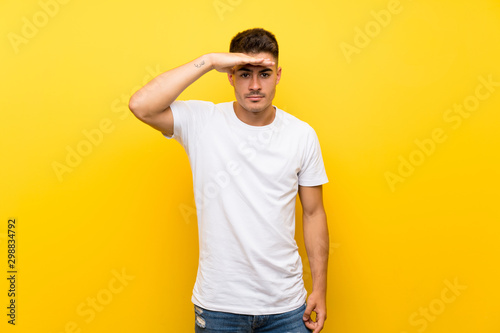 Young handsome man over isolated yellow background looking far away with hand to look something