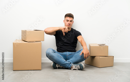 Handsome young man moving in new home among boxes showing thumb down sign