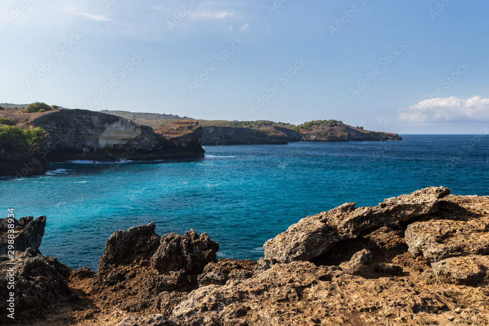 Coastal landscape at Broken Beach, Nusa Pendia, Indonesia. Rocks in foreground showing the rough terrain; clear blue green water below the cliff. Rugged coastline in the distance; blue sky and clouds Coastal landscape at Broken Beach, Nusa Pendia, Indonesia. Rocks in foreground showing the rough terrain; clear blue green water below the cliff. Rugged coastline in the distance; blue sky and clouds