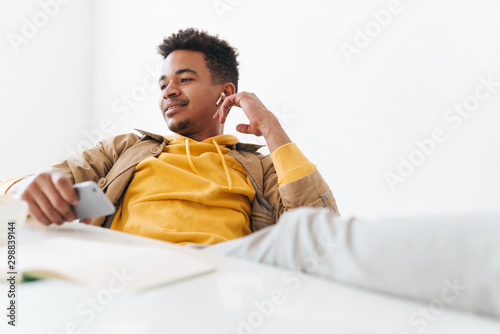 Image of african american guy using smartphone and earbuds in office