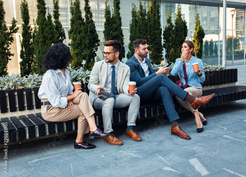 Group of coworkers having a coffee brake together, siting outside in front of office buildings and talking.