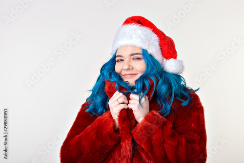 Christmas  fashion and youth - Beautiful young woman with blue hair in red fur coat and santa hat  looking at the camera and standing against a white background. Ready for party.