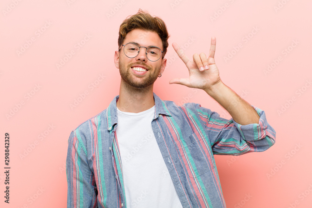 young man feeling happy, fun, confident, positive and rebellious, making rock or heavy metal sign with hand against pink background