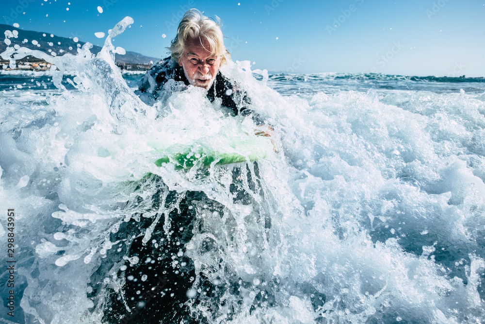 senior trying to surf a wave on the sea at the beach alone with black wetsuit and green surftable - vacation at the sea and active retired man