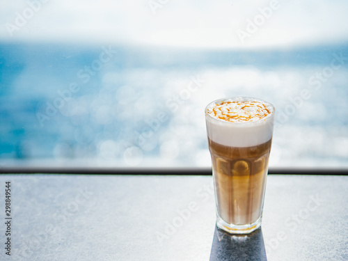 Hot latte macchiato coffee with caramel on tabletop with blue sea on background. Natural hard daylight. Copy space for text or design photo