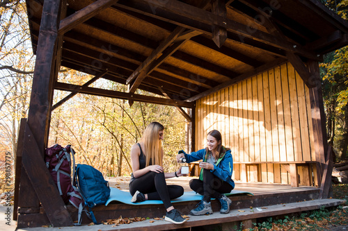 Two girls in sportswear sitting in woods under canopy and drink tea from thermos