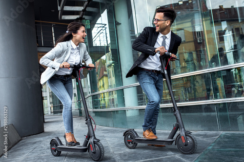Two smiling business people driving electric scooter in front of modern business building going on work. photo