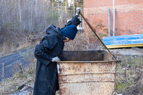 homeless woman in black coat and blue hat is open street garbage cans. woman is rummaging for some food in garbage. poverty.