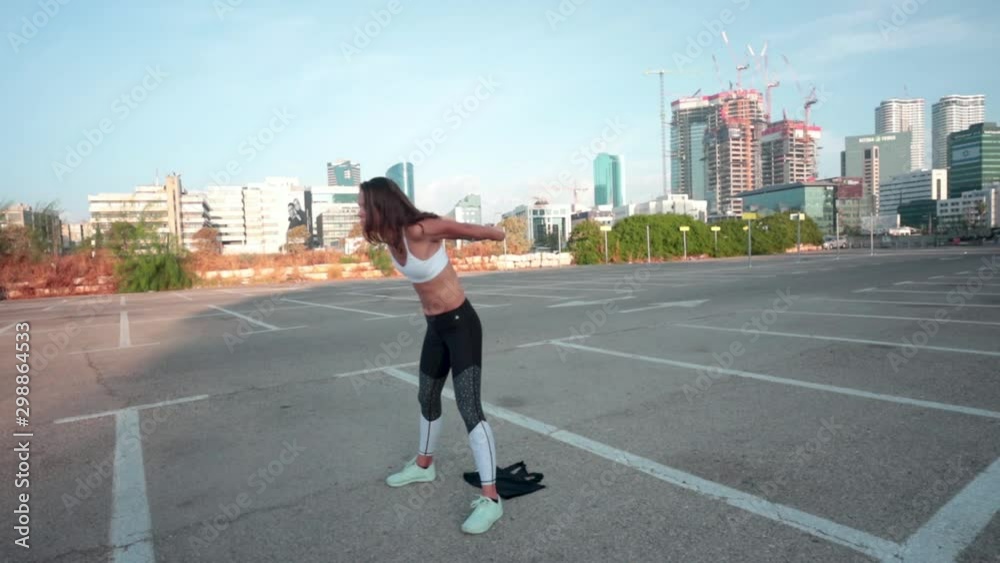 Thin muscular woman doing warm up exercises before her fitness workout. Slow motion shot in a parking lot with an urban skyline background.