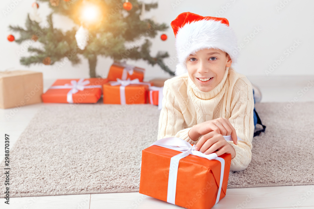 Holidays, christmas, childhood and people concept - smiling happy teen boy in santa hat with gift box over christmas tree background